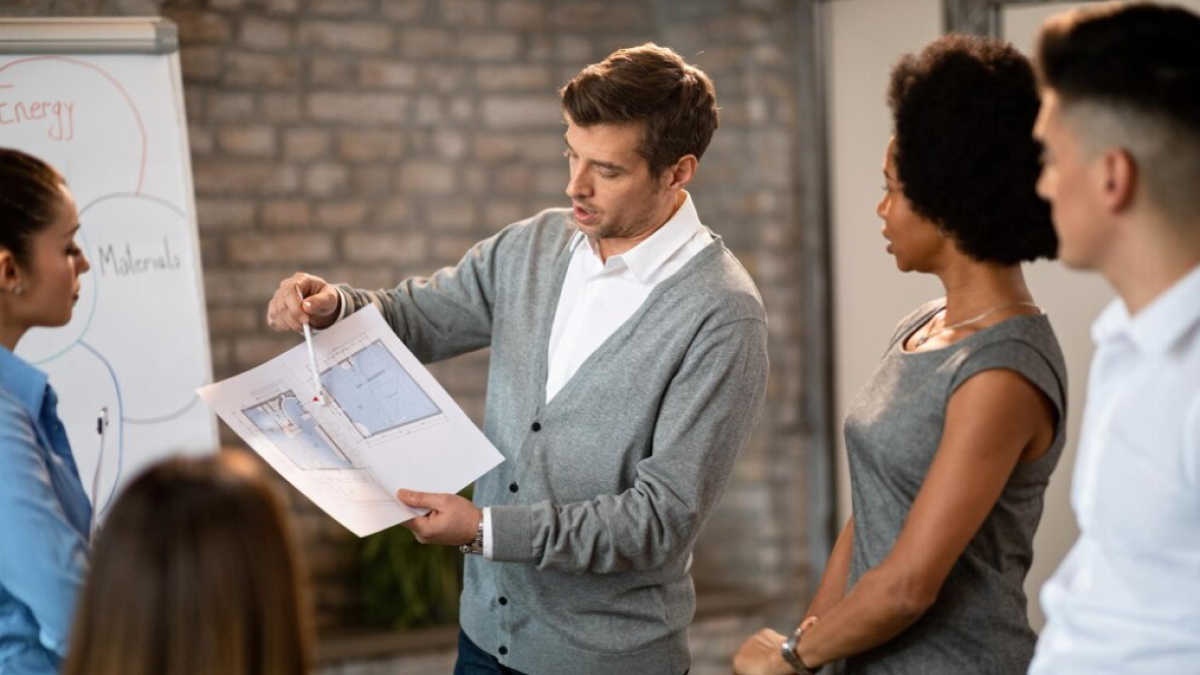 Photo montrant un agent immobilier durant une visite avec un acheteur et une acheteuse. Il montre le plan du logement.