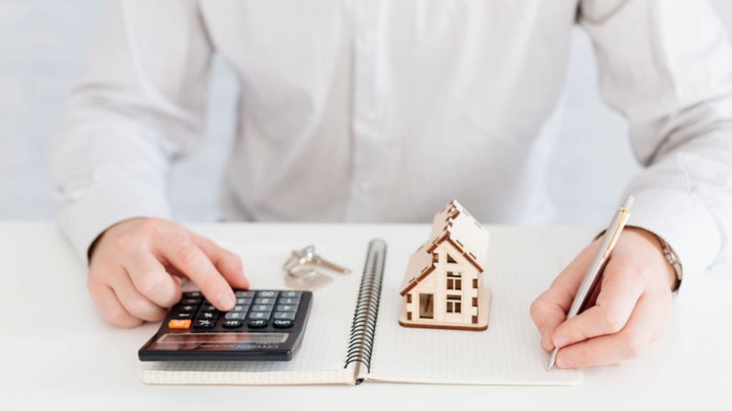 Photo montrant un homme avec une calculatrice, un crayon, des clés, un bloc-note et une petite maison